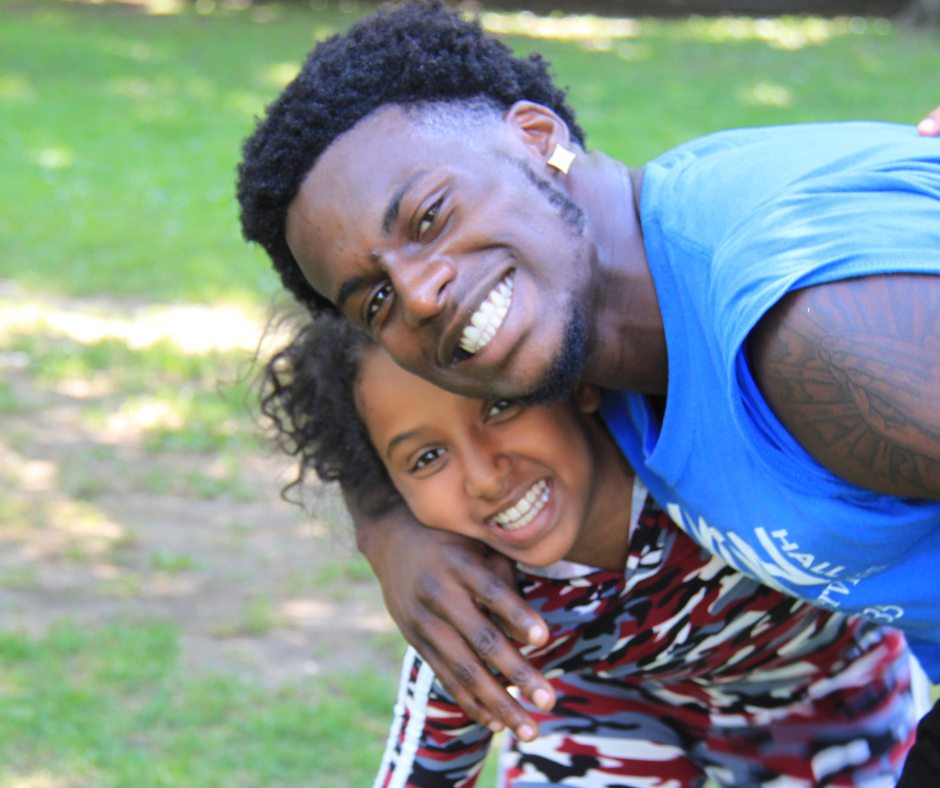 Two people smiling outdoors, with an adult in a blue shirt hugging a child in a patterned outfit. They are both looking at the camera. Grass is visible in the background.