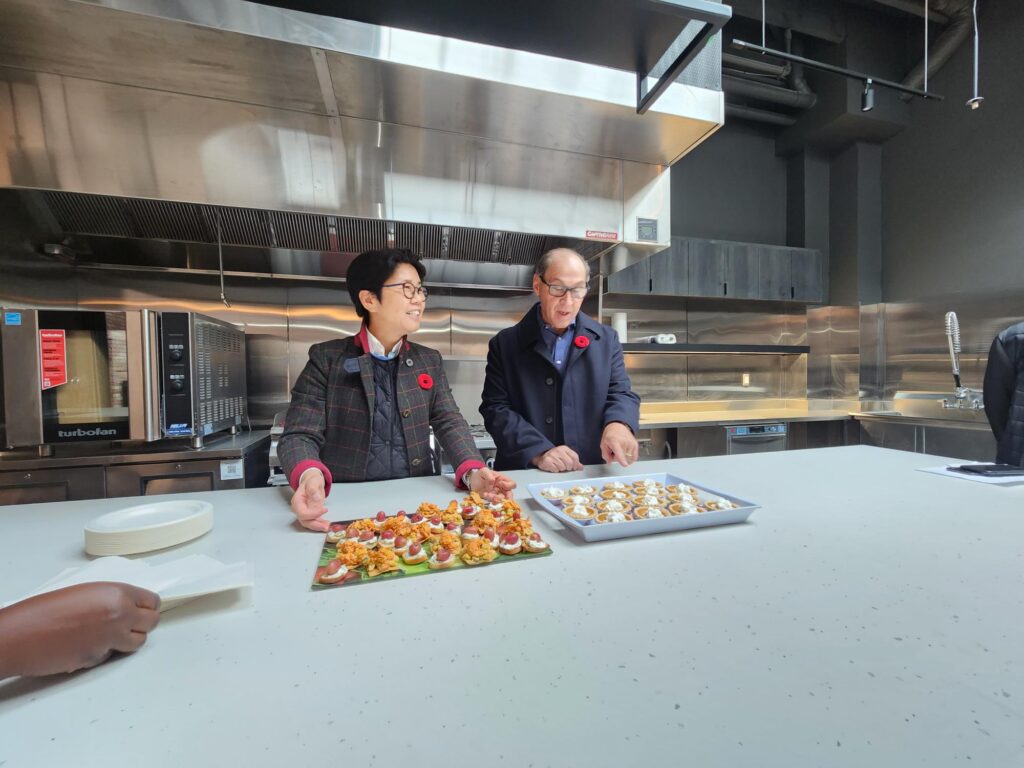 two-people-stand-in-a-modern-kitchen-engaged-in-conversation-while-presenting-trays-of-appetizers-on-a-large-white-countertop-one-tray-holds-colorful-bite-sized-hors-d-oeuvres-while-the-other-has-small-pastries-with-cream-both-individuals-are-wearing-dark-jackets-and-red-poppy-pins-stainless-steel-appliances-and-countertops-are-visible-in-the-background-a-hand-reaching-for-a-plate-is-partially-visible-in-the-foreground-