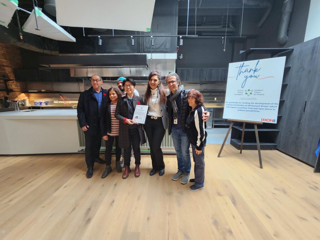a-group-of-six-people-stands-together-in-a-modern-kitchen-space-smiling-for-the-camera-one-person-in-the-center-holds-a-framed-certificate-a-large-thank-you-sign-from-dixon-hall-is-displayed-on-an-easel-in-the-background-acknowledging-support-for-the-kitchen-s-development-the-group-is-dressed-in-a-mix-of-business-and-casual-attire-with-some-wearing-poppy-pins-