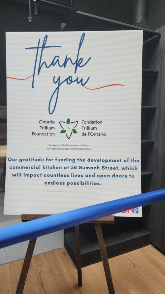 a-large-sign-on-an-easel-displays-a-thank-you-message-from-the-ontario-trillium-foundation-the-text-expresses-gratitude-for-funding-the-development-of-a-commercial-kitchen-at-58-sumach-street-highlighting-its-positive-impact-a-blue-ribbon-is-stretched-across-the-foreground-suggesting-a-ribbon-cutting-ceremony-or-grand-opening-event-