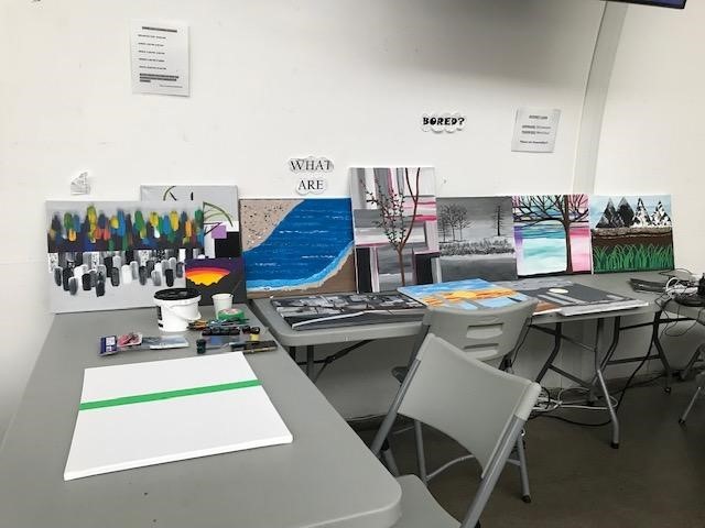 a-table-with-various-paintings-and-art-supplies-is-set-up-in-a-room-with-white-walls-several-colorful-paintings-including-abstract-and-landscape-designs-are-displayed-against-the-wall-a-blank-canvas-with-a-green-stripe-sits-on-the-table-along-with-paintbrushes-markers-and-cups-chairs-are-arranged-around-the-table-suggesting-an-art-activity-or-workshop-signs-on-the-wall-include-the-words-what-are-and-bored