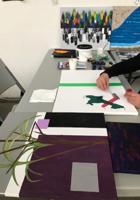 a-person-is-painting-on-a-canvas-at-a-table-filled-with-art-supplies-the-canvas-has-a-green-stripe-and-an-abstract-design-in-progress-other-completed-paintings-featuring-abstract-and-landscape-elements-are-displayed-on-the-table-and-against-the-wall-a-cup-of-water-and-paintbrushes-are-also-present-indicating-an-active-art-session-