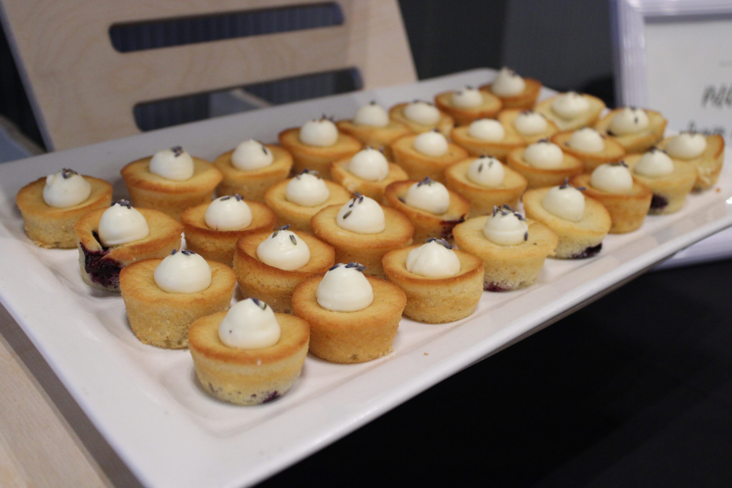 A white platter filled with small, golden-brown mini cakes topped with dollops of white cream and garnished with tiny lavender sprigs.