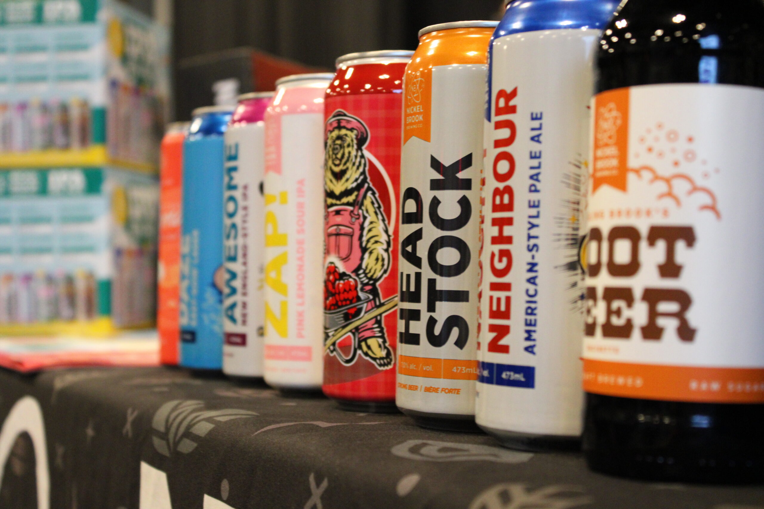 A row of colorful beverage cans, including craft beers and root beer, displayed on a table with a blurred background of stacked boxes.