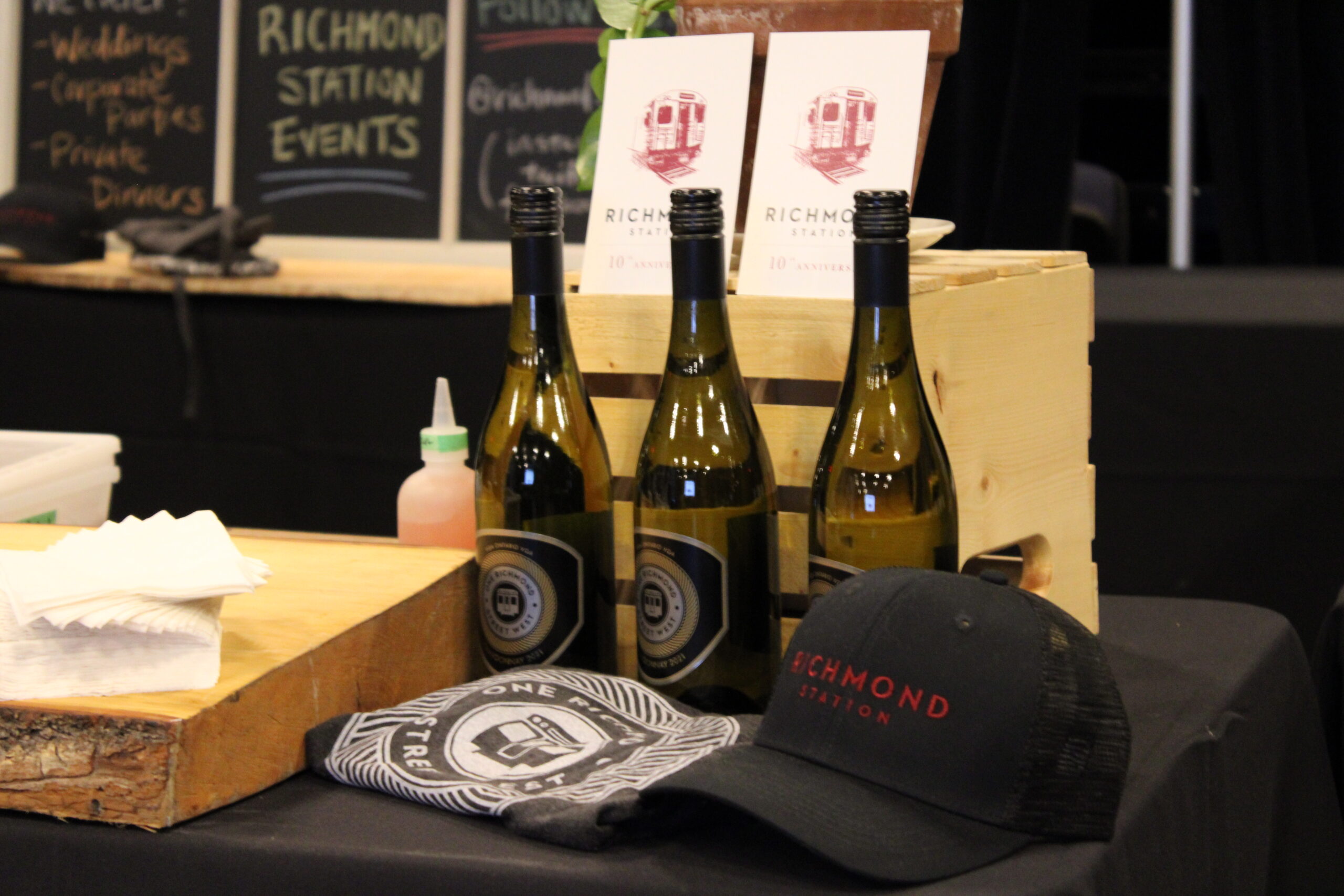 Richmond Station promotional setup featuring three bottles of wine, branded merchandise including a black cap and a folded t-shirt, and wooden crates with event flyers.