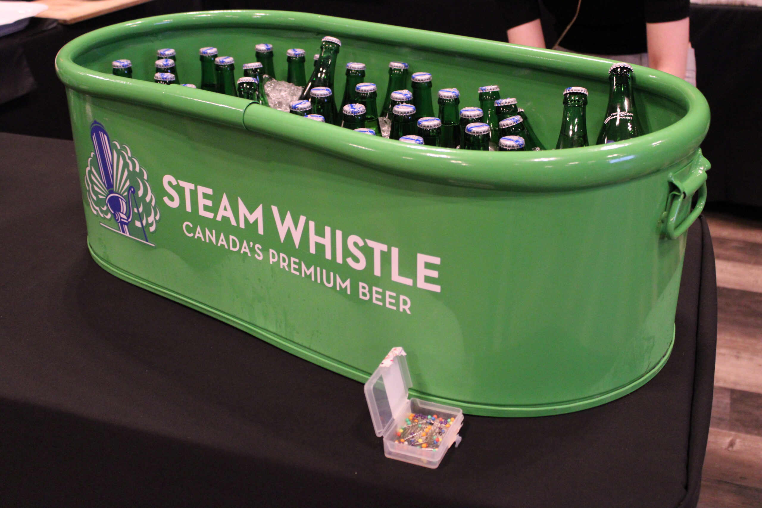 A green Steam Whistle beer cooler filled with ice and bottled beers, placed on a black tablecloth. A small open container of colorful push pins is in the foreground.