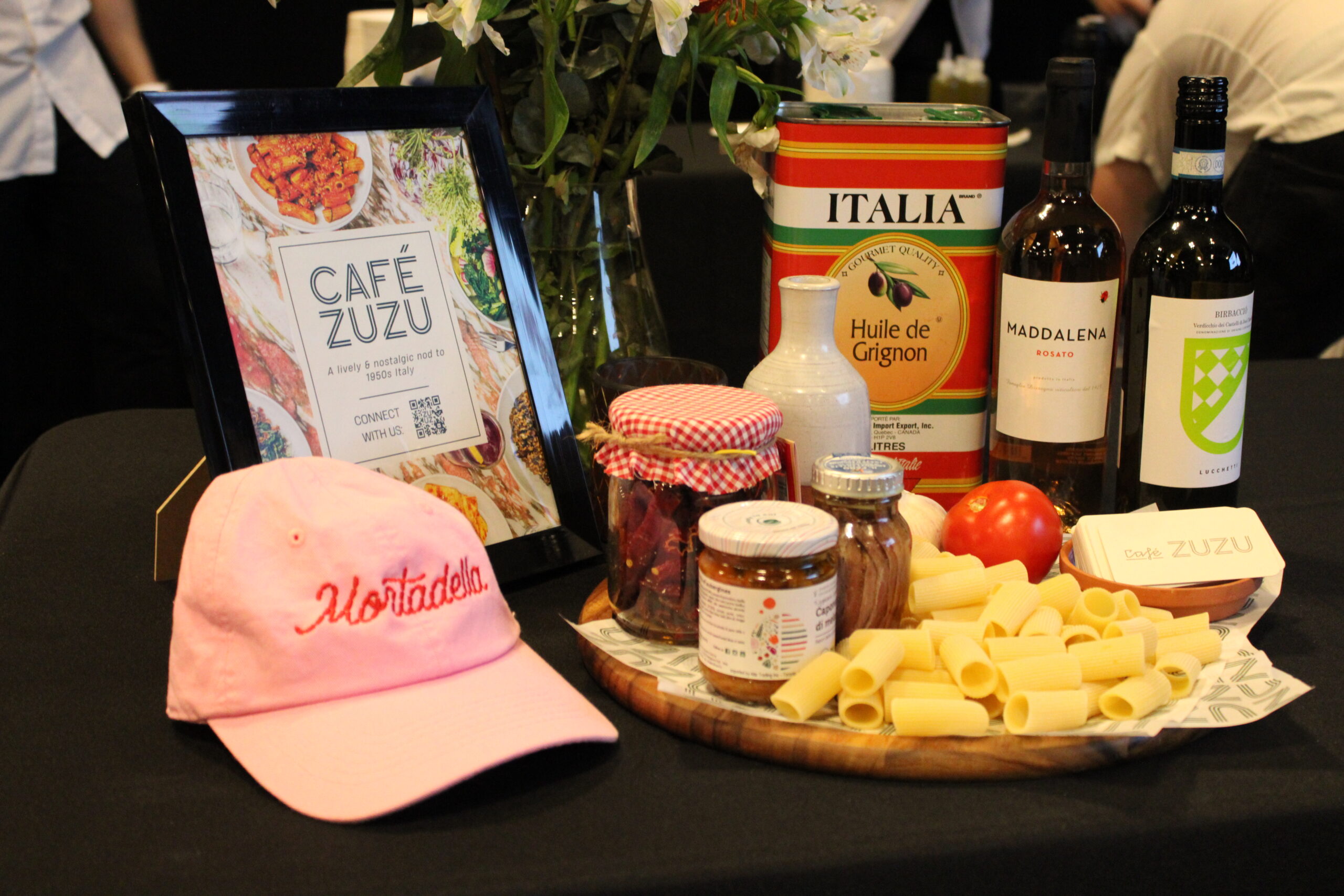 A Café Zuzu promotional display on a black tablecloth, featuring a framed sign, a pink hat embroidered with 'Mortadella,' jars of ingredients, a wooden board with uncooked pasta, tomatoes, bottles of olive oil and wine, and a stack of business cards.