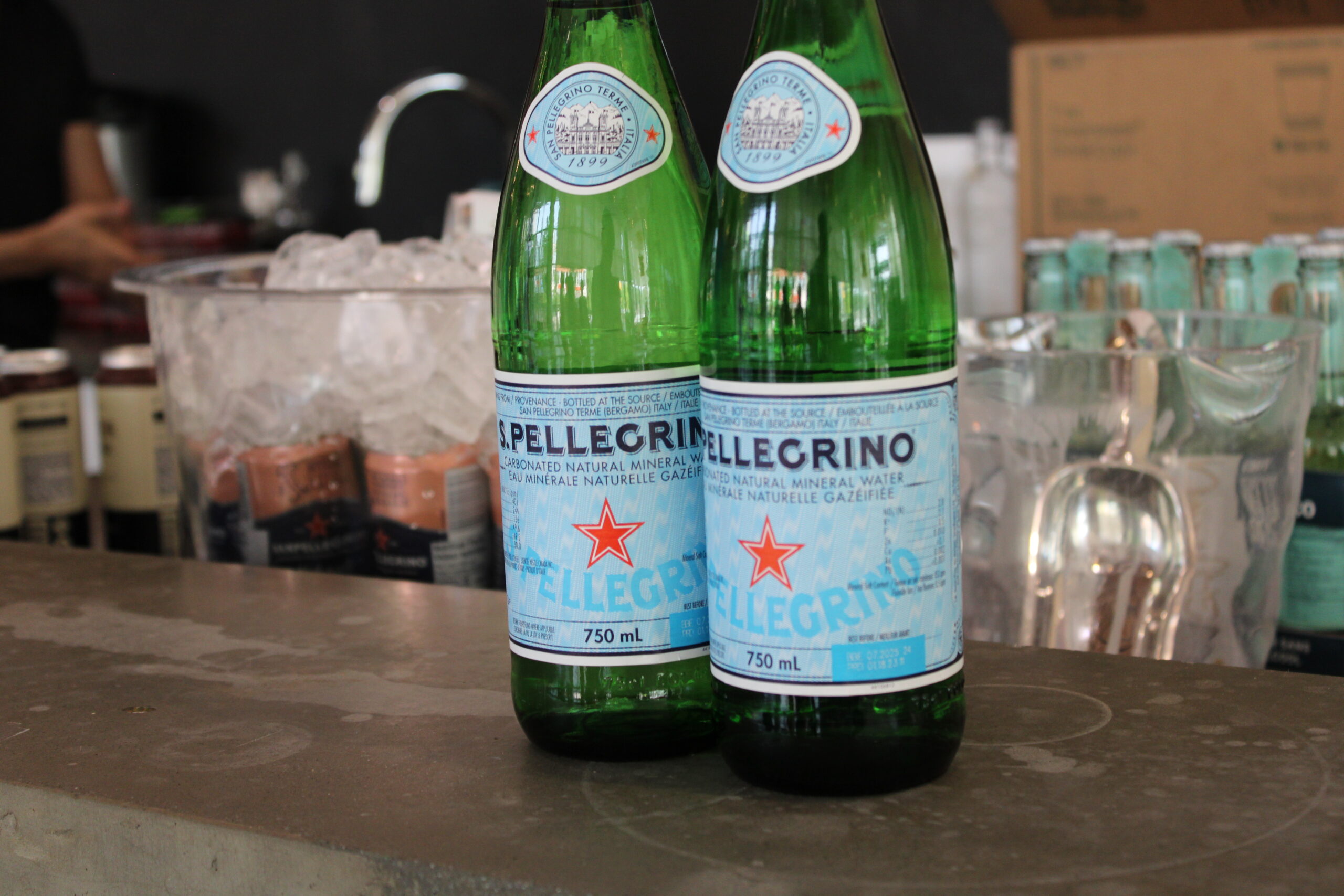 Two bottles of S.Pellegrino sparkling water on a countertop, with a background featuring an ice bucket filled with canned beverages, glassware, and a person working behind the counter.