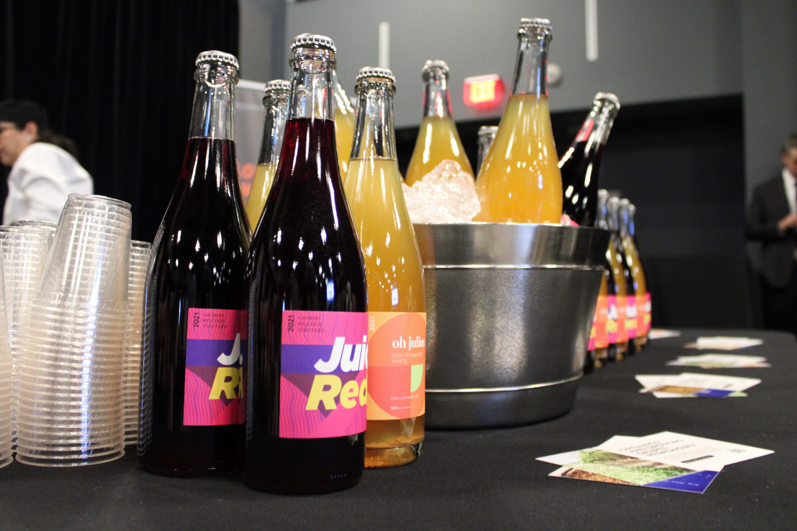 Assorted bottles of red and white beverages displayed on a table, with some chilling in an ice bucket. Stacks of clear plastic cups are nearby, and promotional cards are spread across the table. A bartender and a man in a suit are visible in the background.