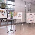 An indoor art auction display featuring framed and unframed artwork on easels, along with a thank-you board listing auction donors. Large windows provide a view of the outside.