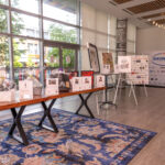An indoor auction display featuring gift baskets, framed artwork on easels, and a thank-you board for donors. Large windows allow natural light to enter the space.