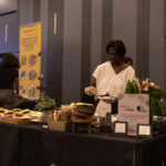 A woman in a white blouse serves food at a catering table filled with sandwiches, fresh ingredients, and promotional signage. A yellow "FoodSquad" banner is visible in the background.