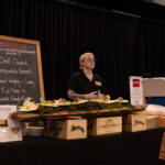 A person in a black shirt stands behind a food display at an event, with plated dishes arranged on a rustic wooden board. A chalkboard sign with "HOOKED INC" and event details is visible in the background.