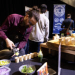 A man wearing glasses and an apron pours a liquid into small cups at a food event. A table with fresh ingredients and a "GLB" brewery banner are visible in the background.