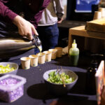 A close-up of a person pouring a liquid into small paper cups at a food event. Fresh ingredients, including chopped vegetables and herbs, are arranged on the table.