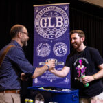 Two men shake hands and smile at a Great Lakes Brewery booth. One man holds a canned beverage, while a large GLB banner is displayed in the background.