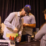 A man wearing a black cap pours a yellow beverage from a pitcher into a cup at a tasting event. Bottles of wine and a sign reading "Last Straw" are visible on the table.