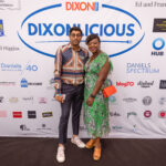 A man and a woman smile and pose together in front of a large "DIXONLICIOUS" event backdrop featuring sponsor logos.