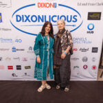 Two women smile and pose together in front of a large "DIXONLICIOUS" event backdrop featuring sponsor logos.