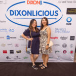 Two women pose and smile in front of a "DIXONLICIOUS" event backdrop featuring various sponsor logos.