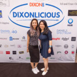 Two women smile and pose together in front of a "DIXONLICIOUS" event backdrop featuring various sponsor logos.