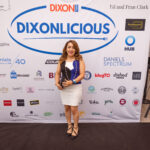 A woman in a stylish black, white, and blue dress poses in front of a "DIXONLICIOUS" event backdrop featuring various sponsor logos.