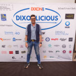 A man in a casual blue outfit with a vest and sunglasses poses in front of a "DIXONLICIOUS" event backdrop featuring various sponsor logos.