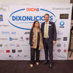 A man in a suit and a woman in a black outfit with a patterned scarf pose in front of a "DIXONLICIOUS" event backdrop featuring various sponsor logos.