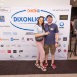 A smiling couple poses in front of a "DIXONLICIOUS" event backdrop featuring various sponsor logos. The woman wears a light blue top and pink pants, while the man wears a navy blue t-shirt and khaki shorts.