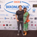 A smiling couple poses in front of a "DIXONLICIOUS" event backdrop featuring various sponsor logos. The man wears a black t-shirt and jeans, while the woman wears a green patterned dress with an orange purse.