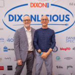 Two men with glasses and smiles pose in front of a "DIXONLICIOUS" event backdrop featuring various sponsor logos. One wears a blazer and white shirt, while the other wears a navy sweater and gray pants.