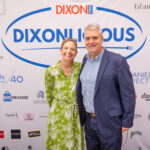 A smiling man and woman pose in front of a "DIXONLICIOUS" event backdrop featuring sponsor logos. The woman wears a green patterned dress, and the man wears a blue blazer over a checkered shirt.