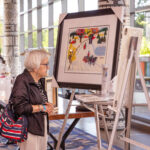 An elderly woman with glasses and a striped bag observes a colorful framed painting on display at an art auction or exhibition.