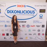 A woman in a black dress poses in front of a "Dixonlicious" event backdrop featuring various sponsor logos.
