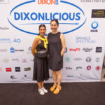 Two women in black dresses pose in front of a "Dixonlicious" event backdrop featuring various sponsor logos. One has a yellow sweater tied around her shoulders.