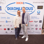 Two men pose in front of a "Dixonlicious" event backdrop featuring various sponsor logos. One is wearing a beige jacket and glasses, while the other is dressed in a blazer and sneakers.