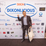 A man in a suit poses in front of a "Dixonlicious" event backdrop, pointing at the logo while holding a tote bag.