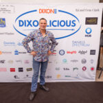 A man in a colorful patterned shirt and jeans poses in front of the "Dixonlicious" event backdrop, smiling with hands on hips.
