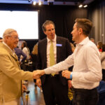Three men engaged in conversation at an event, with two shaking hands while the third observes. The background shows a crowded room with attendees.