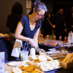 A woman wearing gloves prepares food at a catering station, assembling sandwiches with fresh ingredients. A tray of drinks and event attendees are visible in the background.