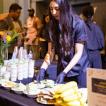 A woman wearing gloves prepares and arranges food plates at a catering station, with stacks of sauce cups, bananas, and event attendees in the background.