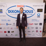 A man in a dark blazer and white shirt stands in front of a "DIXONLICIOUS" event backdrop with various sponsor logos.