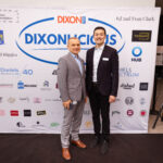 Two men in suits stand in front of a "DIXONLICIOUS" event backdrop with various sponsor logos, smiling at the camera.