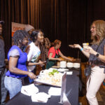 A group of people gathers around a food station, engaging in conversation while sampling dishes. A woman in a gray top holds a drink and a small plate, interacting with the servers behind the table.