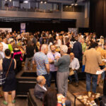 A large crowd gathers at a networking or social event, engaging in conversations and enjoying food and drinks in a spacious venue.