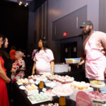Two vendors wearing pink aprons stand behind a dessert table filled with cupcakes and macarons, engaging with attendees at an event.