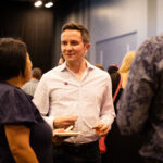 A man in a white patterned shirt holds a drink and a small plate while engaged in conversation at a social event.
