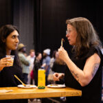 Two women stand at a high-top table, enjoying food and drinks while engaged in conversation at a social event.