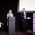 A woman stands at a podium speaking while a man beside her holds a microphone and papers, addressing an audience at an event.