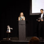 A woman stands at a podium, speaking and clapping, while a man beside her holds papers and a microphone on a stage at an event.